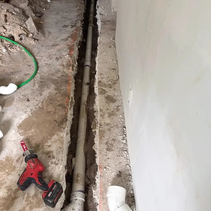 A drain line exposed in a trench cut through a concrete slab inside a house, with a cordless drill on the floor beside it